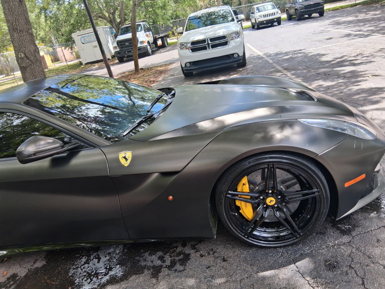 Matte black Ferrari sports car with yellow brake calipers parked in a sunny paved parking lot.