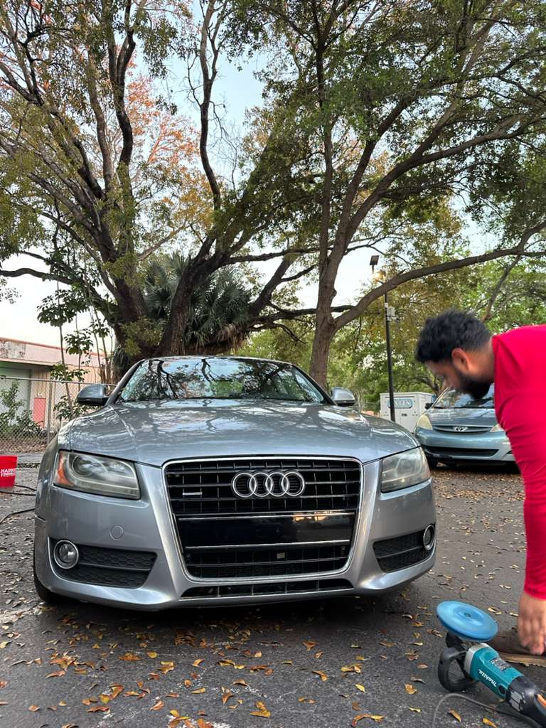 Man in red shirt polishes a silver Audi car with a power buffer outdoors.
